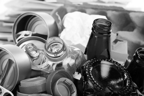 A collection of various discarded waste items placed on a surface, including several empty glass and plastic bottles, with some still having caps attached or nearby, and a few metal cans and containers visible among the debris. The bottles and cans are of different sizes and shapes, with transparent, green, and dark glass or plastic textures, some showing ridges or smooth finishes. In the background, cardboard boxes and crumpled paper are partially visible, suggesting a recycling or rubbish collection scene possibly set on a pavement or driveway in an urban area, consistent with rubbish removal services such as those offered by Flat Clearance Marylebone. The lighting indicates a natural daytime environment, with shadows and reflections on the metallic and glass surfaces, enhancing the cluttered presentation of waste materials. The image illustrates typical refuse that might be targeted for collection and disposal by rubbish clearance specialists operating within central London postcode areas, such as those in Marylebone or nearby districts.