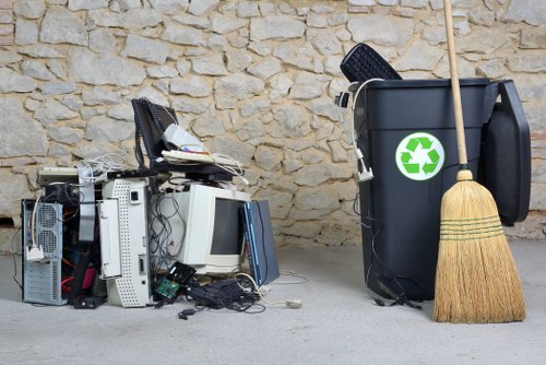 The image displays a collection of discarded electronic waste, including old computer monitors, CPU towers, keyboards, and various cables, all placed on a concrete floor against a textured stone wall. To the right, there is a large black rubbish bin with a green recycling symbol on the front, partially filled with miscellaneous items such as a black plastic bag and a small tool or electronic device resting on top. A wooden broom with a straw head leans against the bin, positioned on the floor beside it. The scene appears to be an outdoor or semi-outdoor area typical for waste collection in a residential or commercial setting in Marylebone, within the postcode area, with natural lighting highlighting the textures of the stone wall and the different materials of the waste items. This setup suggests a focus on rubbish disposal and clearance services provided by Flat Clearance Marylebone, specialising in electronic and general waste removal in the local area.