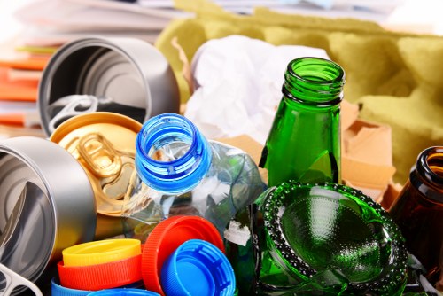 A close-up view of various plastic waste items and discarded bottles, including a green glass bottle and a transparent plastic bottle with a blue cap, all scattered among colorful bottle caps and lids on a surface. In the background, there are crumpled paper, cardboard, and other rubbish, with some foam and packaging materials visible. The scene is indicative of waste collected for rubbish removal, typical of services offered by Flat Clearance Marylebone. The environment appears to be an indoor or courtyard area, suggesting preparing waste for collection or disposal in central London. The textures of plastic, glass, and paper are clearly visible, with the bottles showing smooth and glossy finishes, while the cardboard has a rougher matte texture. The lighting suggests natural ambient light, highlighting the diverse materials. This imagery emphasizes the need for professional rubbish clearance services in Marylebone and nearby areas.