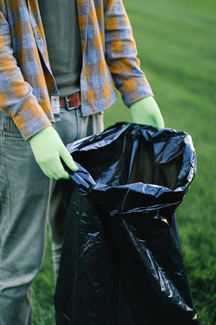 A collection of tightly bundled and crumpled black and white plastic trash bags stacked on a concrete sidewalk in front of a dark metal fence, with trees visible behind the fence. The bags appear fill